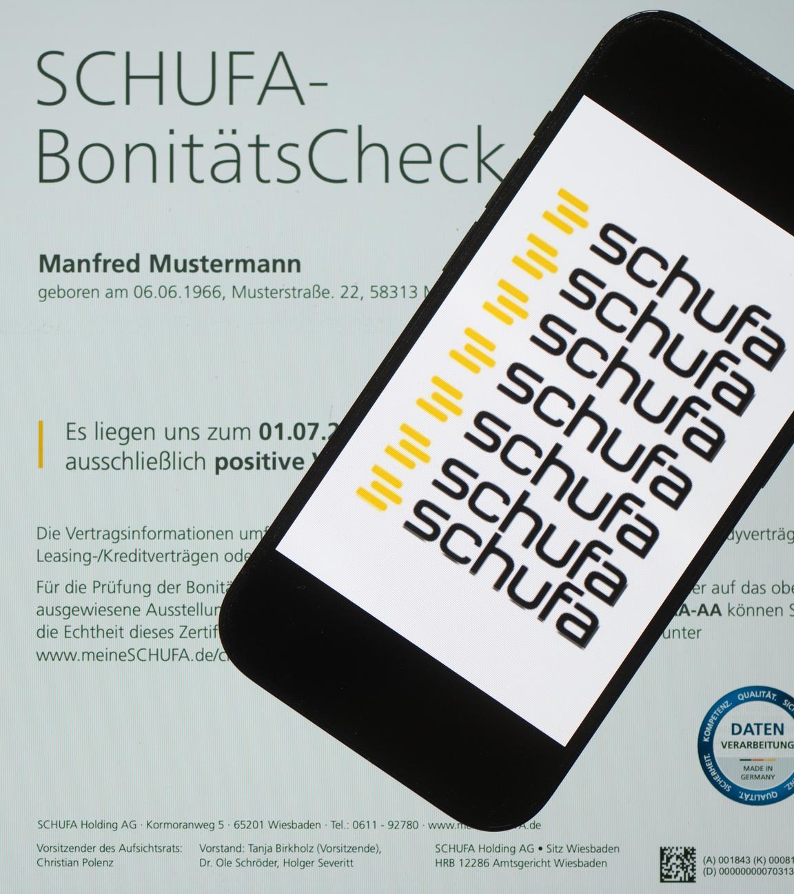 Ein Smartphone mit dem Logo der Schufa liegt auf einem Display mit einem Musterdokument eines Schufa-Bonitätszertifikats.