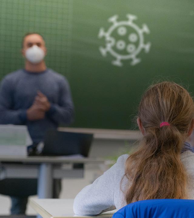 Ein Klassenzimmer mit einem Lehrer mit Maske, der an der Tafel steht, auf die das Corona-Symbol gezeichnet wurde. Im Vordergrund sieht man Kinder