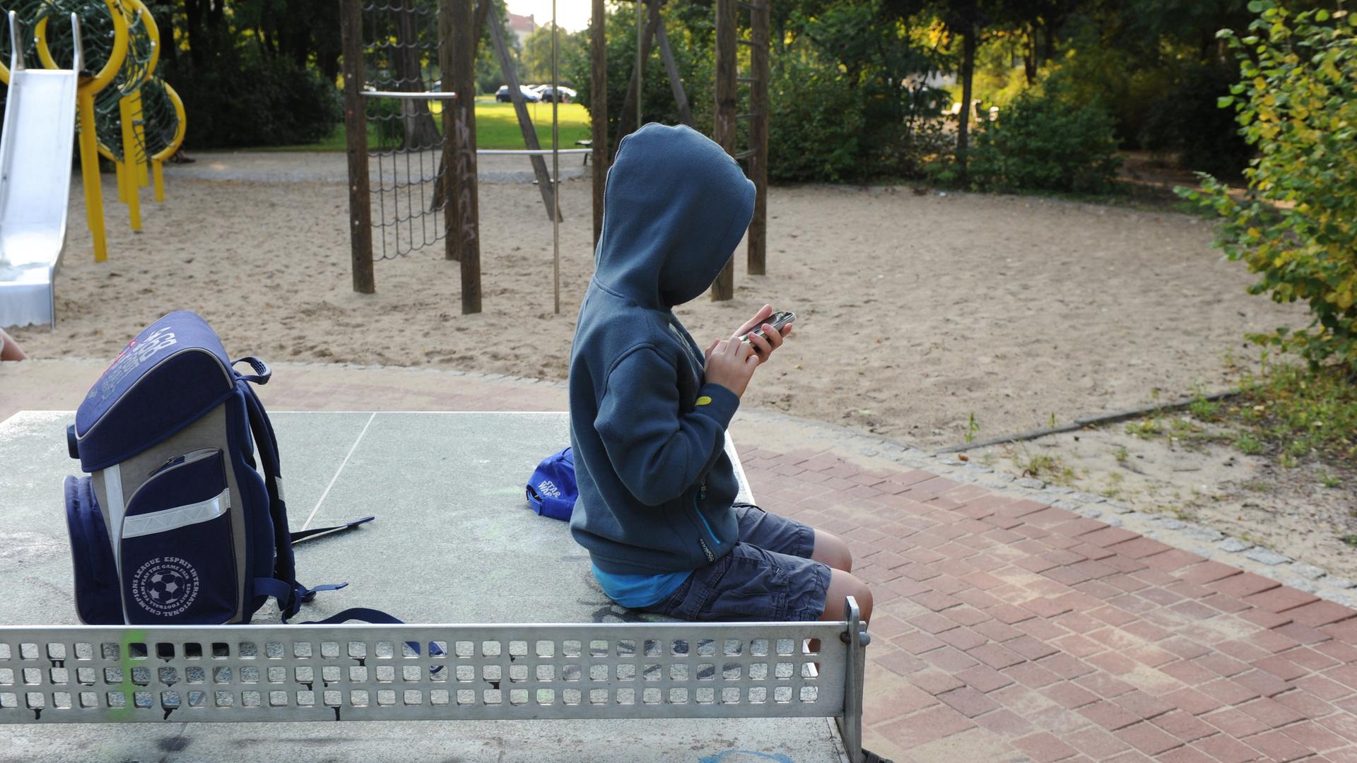 Ein Kind sitzt auf einer Tischtennisplatte auf einem Spielplatz und spielt mit einem Smartphone.