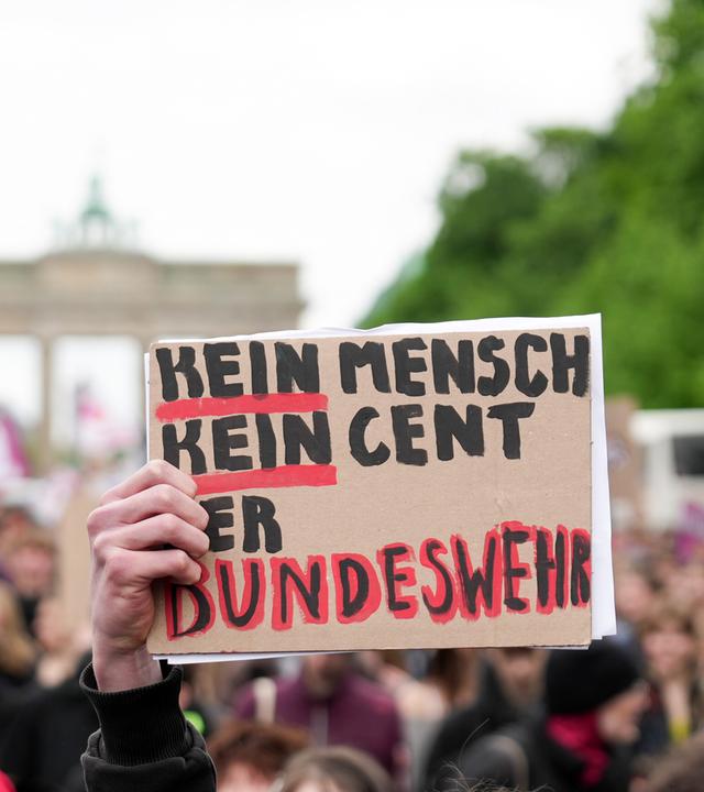 Eine Hand hält ein Schild mit der Aufschrift "Kein Mensch, kein Cent der Bundeswehr" während zahlreiche Demonstranten, am 08.05.2026, in Berlin gegen die Wehrpflicht protestieren.
