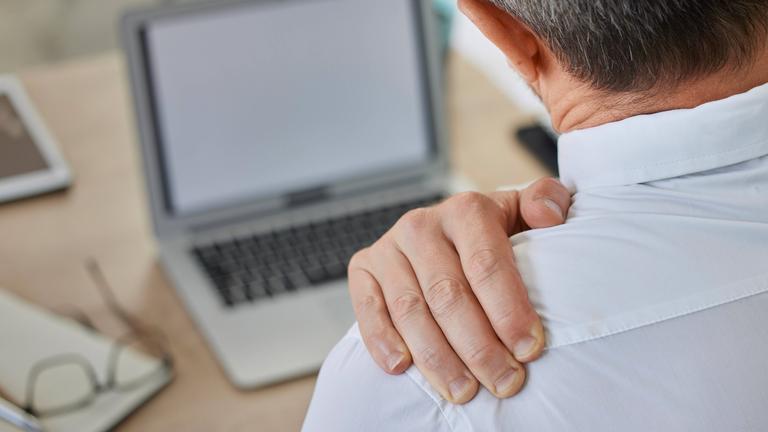 Ein Mann am Schreibtisch mit einem Laptop ist von hinten zu sehen. Er hält sich mit einer Hand die Schulter.