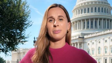 Die ZDF-Korrespondentin in Washington D.C., Katharina Schuster, steht vor dem US-Kongressgebäude. Sie trägt eine dunkle Hose und ein farbiges Oberteil mit freien Armen. An ihrem linken Handgelenk trägt sie eine dunkle Armbanduhr. Mittig auf ihrem Oberteil steckt ein dunkles Mikrofon mit einem flauschigen Windschutz. Zwischen ihr und dem Kongressgebäude ist ein Grünstreifen. Sie blickt direkt in die Kamera. Ihre hellbraunen Haare fallen über die Schultern.