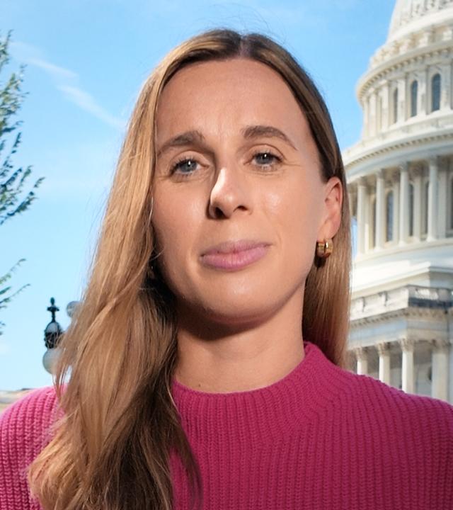 Die ZDF-Korrespondentin in Washington D.C., Katharina Schuster, steht vor dem US-Kongressgebäude. Sie trägt eine dunkle Hose und ein farbiges Oberteil mit freien Armen. An ihrem linken Handgelenk trägt sie eine dunkle Armbanduhr. Mittig auf ihrem Oberteil steckt ein dunkles Mikrofon mit einem flauschigen Windschutz. Zwischen ihr und dem Kongressgebäude ist ein Grünstreifen. Sie blickt direkt in die Kamera. Ihre hellbraunen Haare fallen über die Schultern.