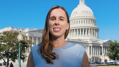 US-Korrespondentin Katharina Schuster steht in einem blauen Oberteil mit kurzen Ärmeln auf dem Rasen vor dem US-Kongressgebäude in Washington DC und spricht in die Kamera. Ihre langen braunen Haare fallen ihr über die rechte Schulter nach vorne. Sie trägt eine dunkle Hose.