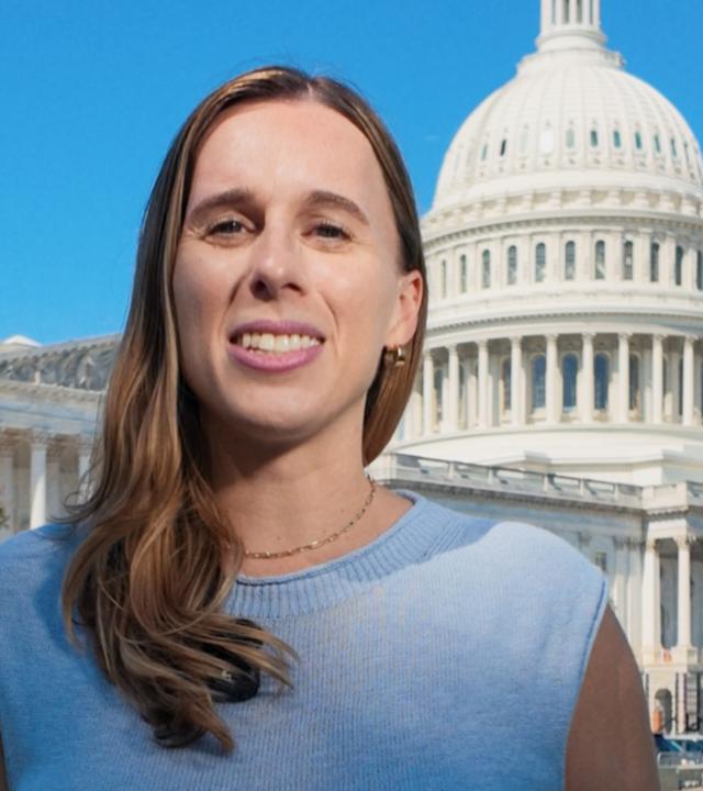 US-Korrespondentin Katharina Schuster steht in einem blauen Oberteil mit kurzen Ärmeln auf dem Rasen vor dem US-Kongressgebäude in Washington DC und spricht in die Kamera. Ihre langen braunen Haare fallen ihr über die rechte Schulter nach vorne. Sie trägt eine dunkle Hose.