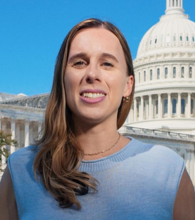 US-Korrespondentin Katharina Schuster steht in einem blauen Oberteil mit kurzen Ärmeln auf dem Rasen vor dem US-Kongressgebäude in Washington DC und spricht in die Kamera. Ihre langen braunen Haare fallen ihr über die rechte Schulter nach vorne. Sie trägt eine dunkle Hose.