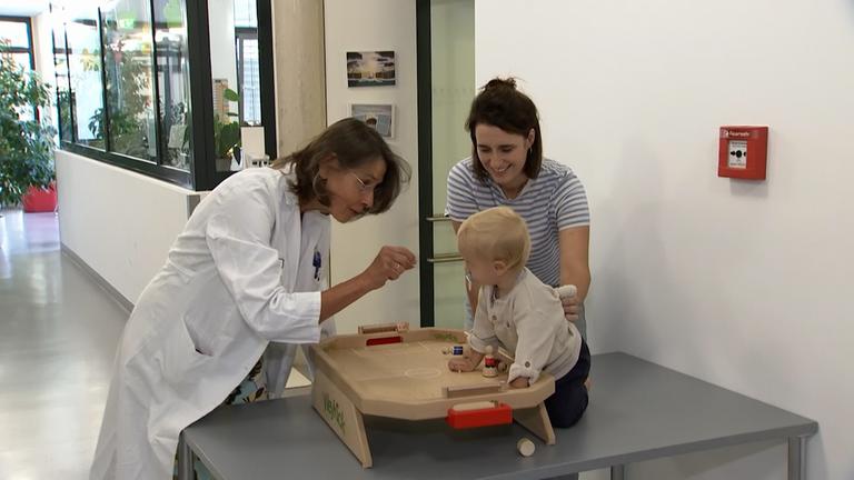 Man sieht eine Mutter die ihr Kind glücklich hält und eine Ärztin, die mit dem Kind spielt.