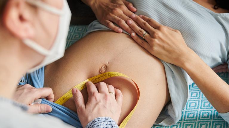 Eine Hebamme misst mit einem Maßband den Symphysen-Fundus-Abstand einer schwangeren Frau.