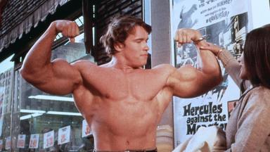 Ein Bodybuilder posiert mit nacktem Oberkörper, während eine Frau ihn am Arm fasst.
