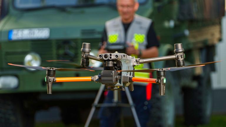 Vor einem Militärauto steht ein Mann mit Fernbedienung, mit der er seine Drohne lenkt.