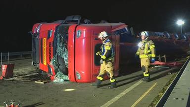 Das Bild zeigt: Umgekippter roter Schwertransporter liegt nachts auf einer Fahrbahn. Zwei Einsatzkräfte in Schutzkleidung gehen an dem beschädigten Fahrzeug vorbei. Die Unfallstelle ist ausgeleuchtet.