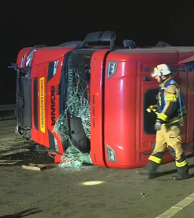 Das Bild zeigt: Umgekippter roter Schwertransporter liegt nachts auf einer Fahrbahn. Zwei Einsatzkräfte in Schutzkleidung gehen an dem beschädigten Fahrzeug vorbei. Die Unfallstelle ist ausgeleuchtet.