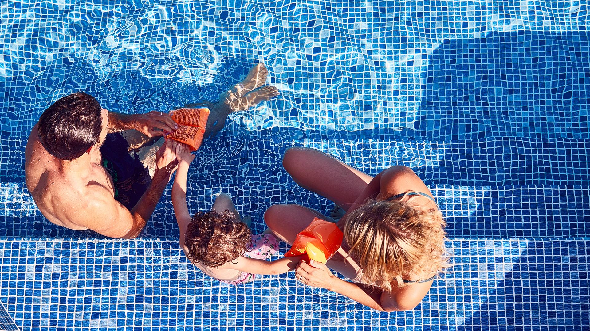 Eltern sitzen auf der Treppe eines Poolbeckens und ziehen ihrem Kind Schwimmflügel an.