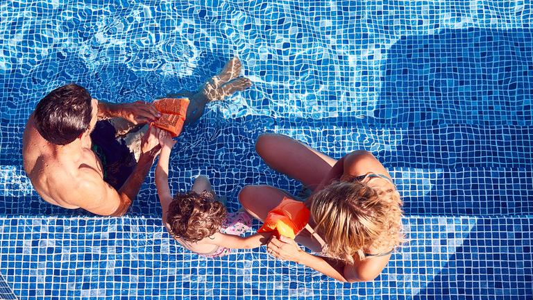 Eltern sitzen auf der Treppe eines Poolbeckens und ziehen ihrem Kind Schwimmflügel an.
