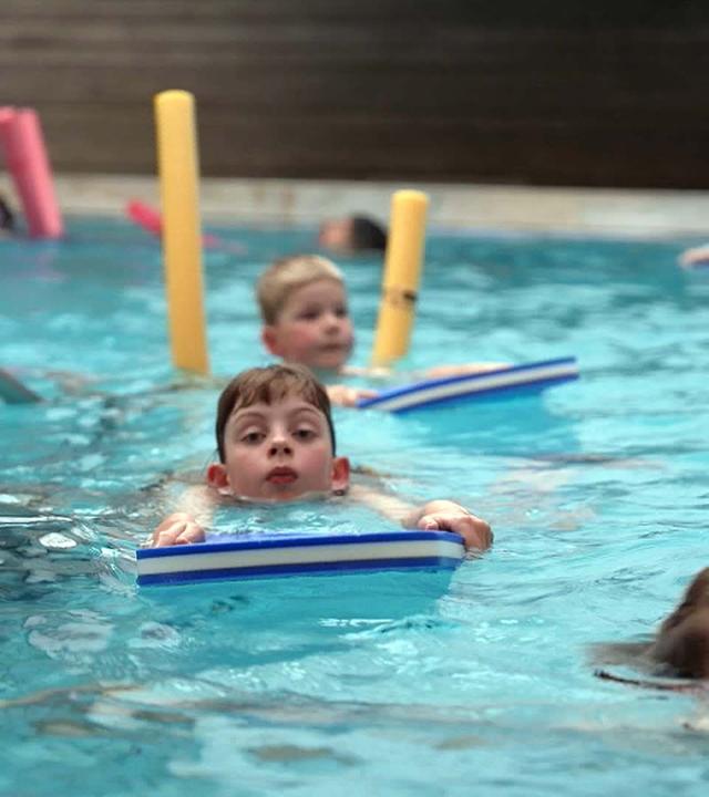 Kinder im Wasser mit Schwimmnudel und -brett beim Schwimmunterricht