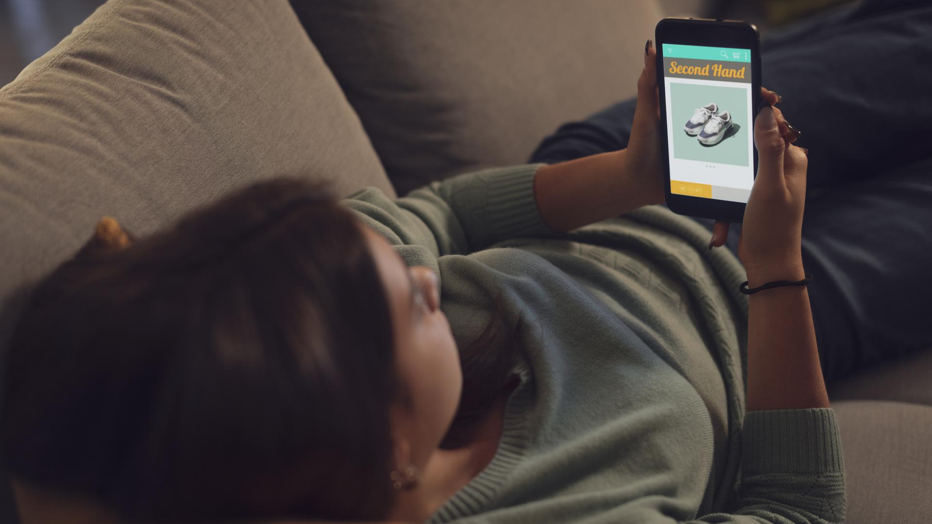 Junge Frau liegt auf dem Sofa und stöbert in den Angeboten einer Secondhand-Shopping-App.