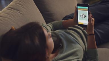 Junge Frau liegt auf dem Sofa und stöbert in den Angeboten einer Secondhand-Shopping-App.