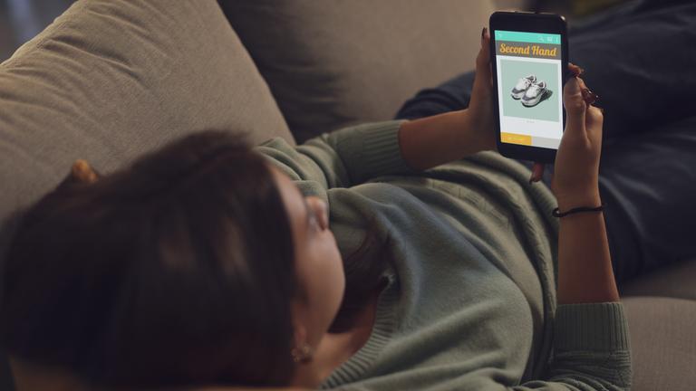 Junge Frau liegt auf dem Sofa und stöbert in den Angeboten einer Secondhand-Shopping-App.