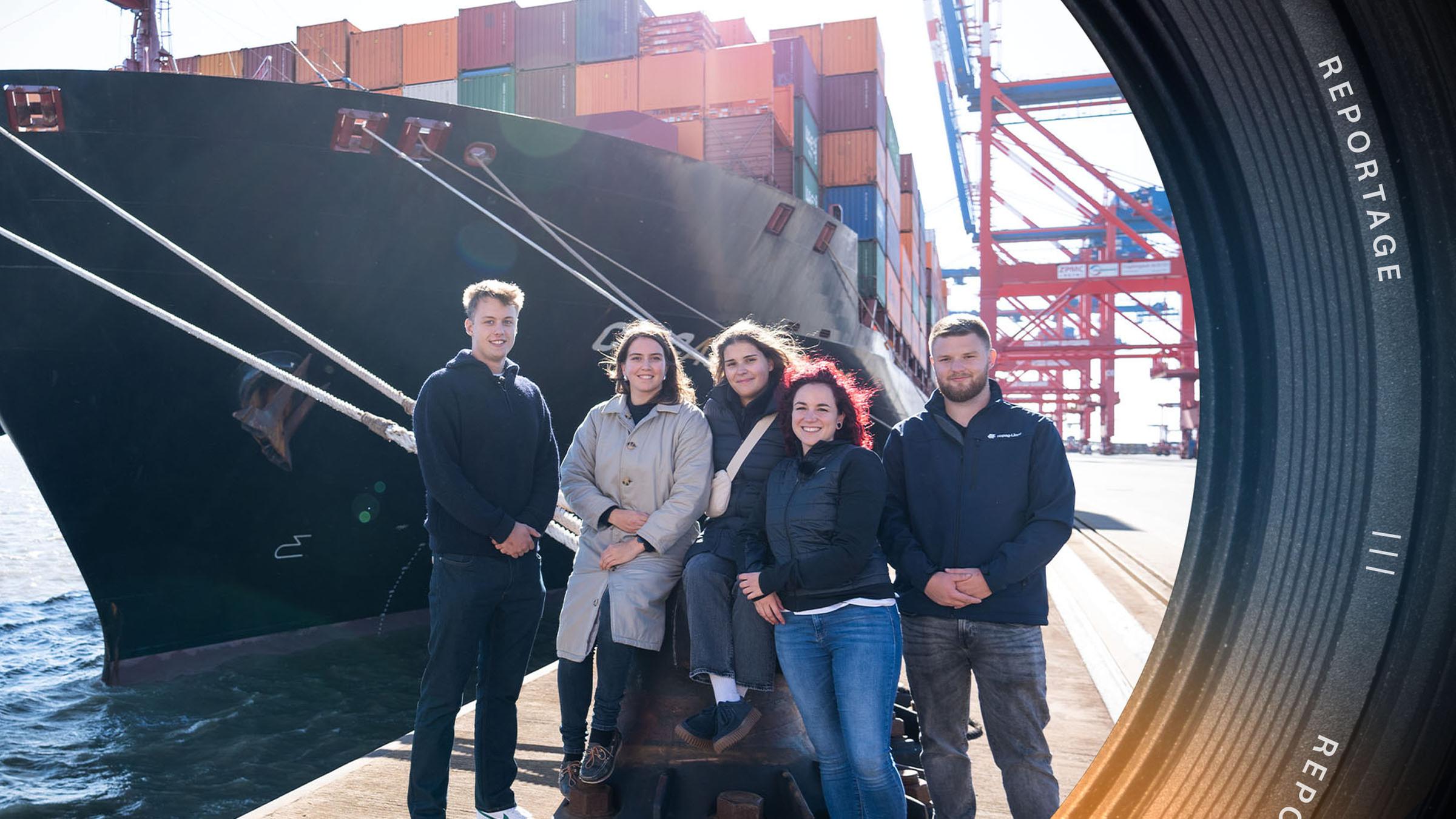 Fünf junge Auszubildende stehen vor einem Containerschiff und schauen in die Kamera.