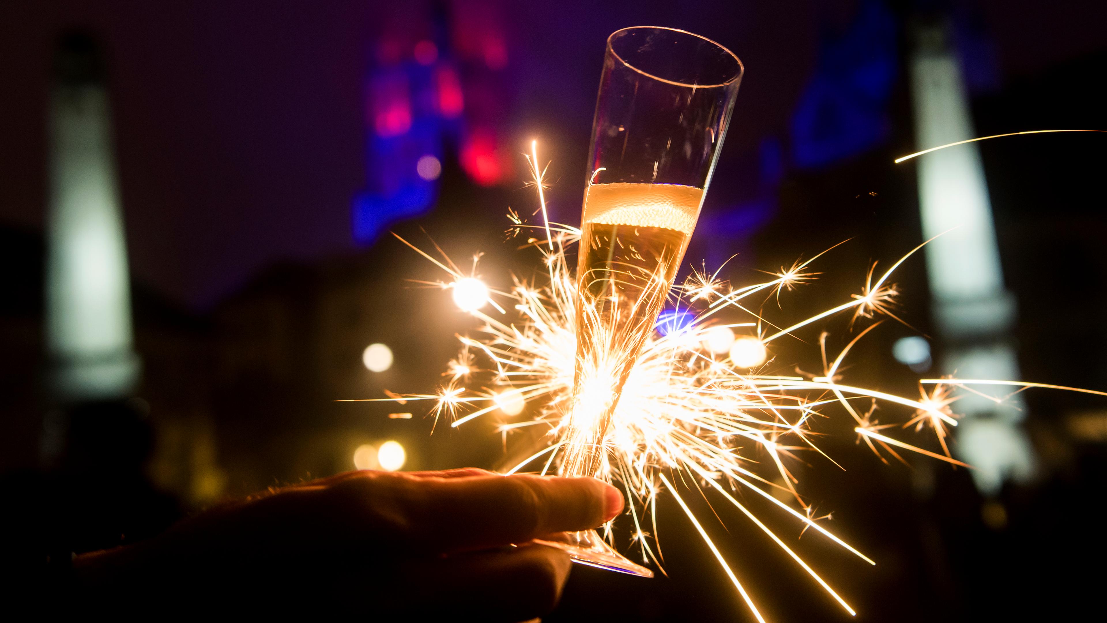 Eine Hand hält ein Glas Sekt in der Hand, dahinter brennt eine Wunderkerze.