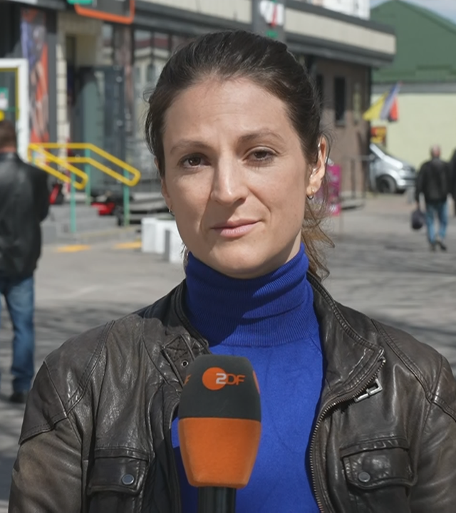 ZDF-Korrespondentin Alica Jung berichtet live aus der Ukraine. Hinter ihr ist bei Sonnenschein eine Fußgängerzone mit vorbeilaufenden Passanten zusehen.