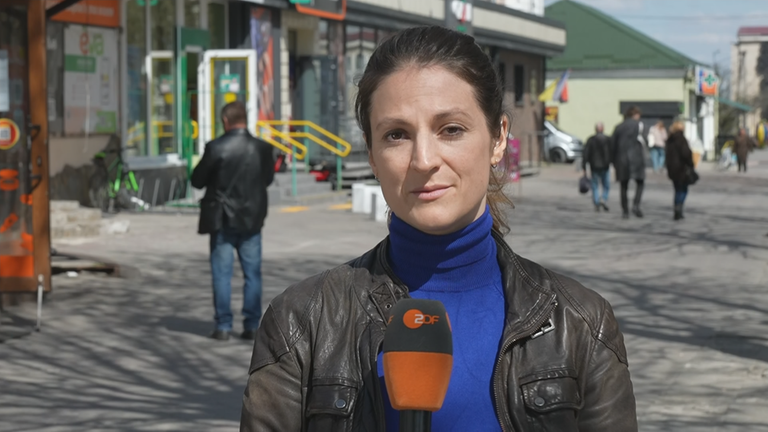 ZDF-Korrespondentin Alica Jung berichtet live aus der Ukraine. Hinter ihr ist bei Sonnenschein eine Fußgängerzone mit vorbeilaufenden Passanten zusehen.