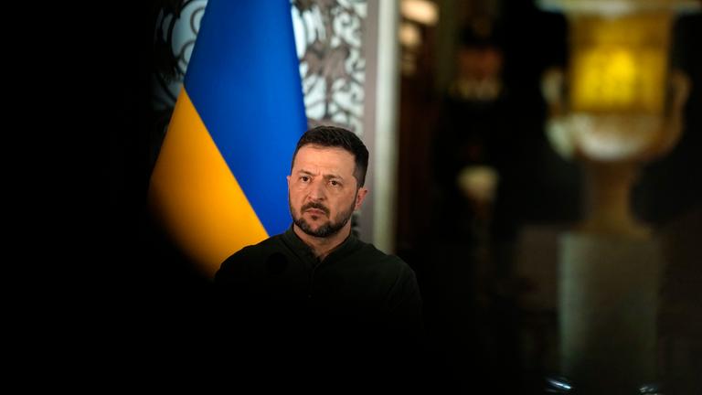 Der Präsident der Ukraine, Wolodymyr Selenskyy steht vor einer ukrainischen Flagge. Um ihn herum ist es dunkel.
