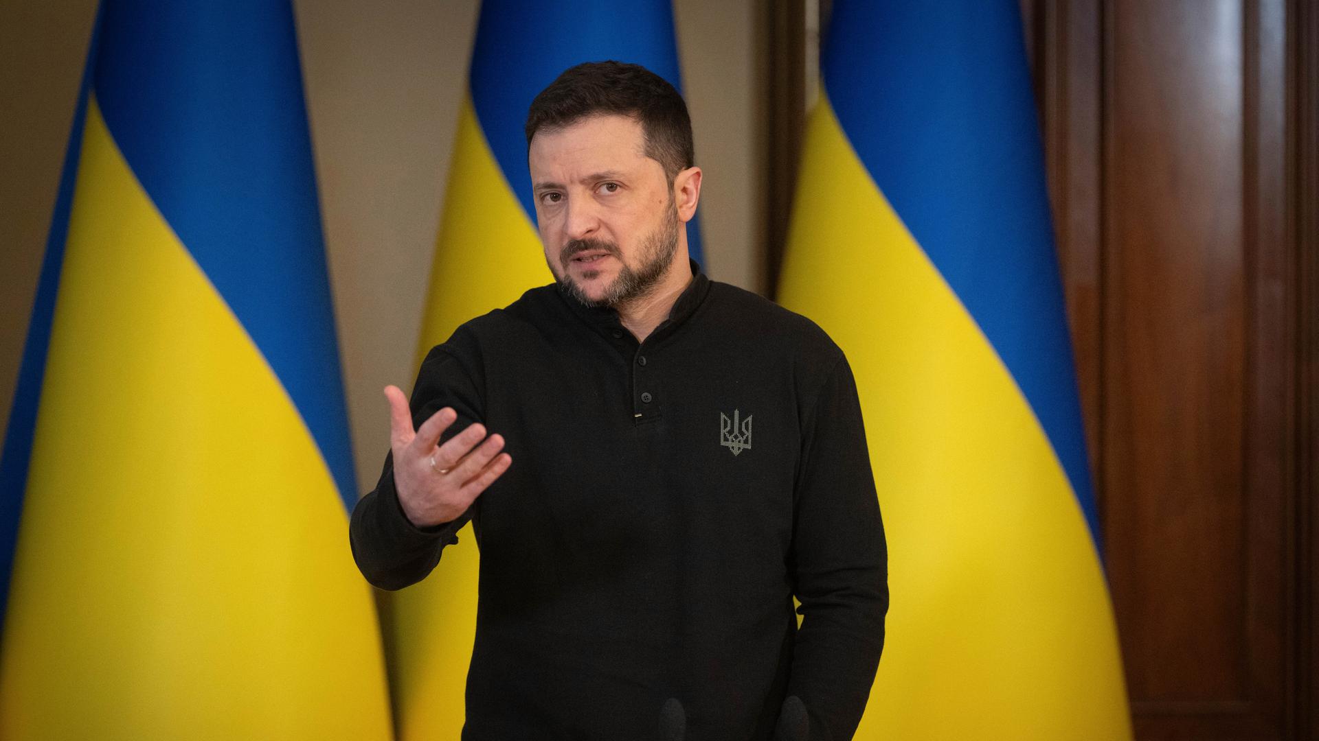 Der ukrainische Präsident Selenskyj der vor drei ukrainischen Flaggen steht und spricht.