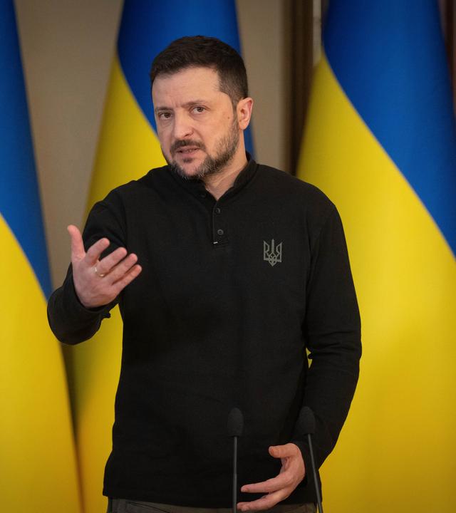 Der ukrainische Präsident Selenskyj der vor drei ukrainischen Flaggen steht und spricht.
