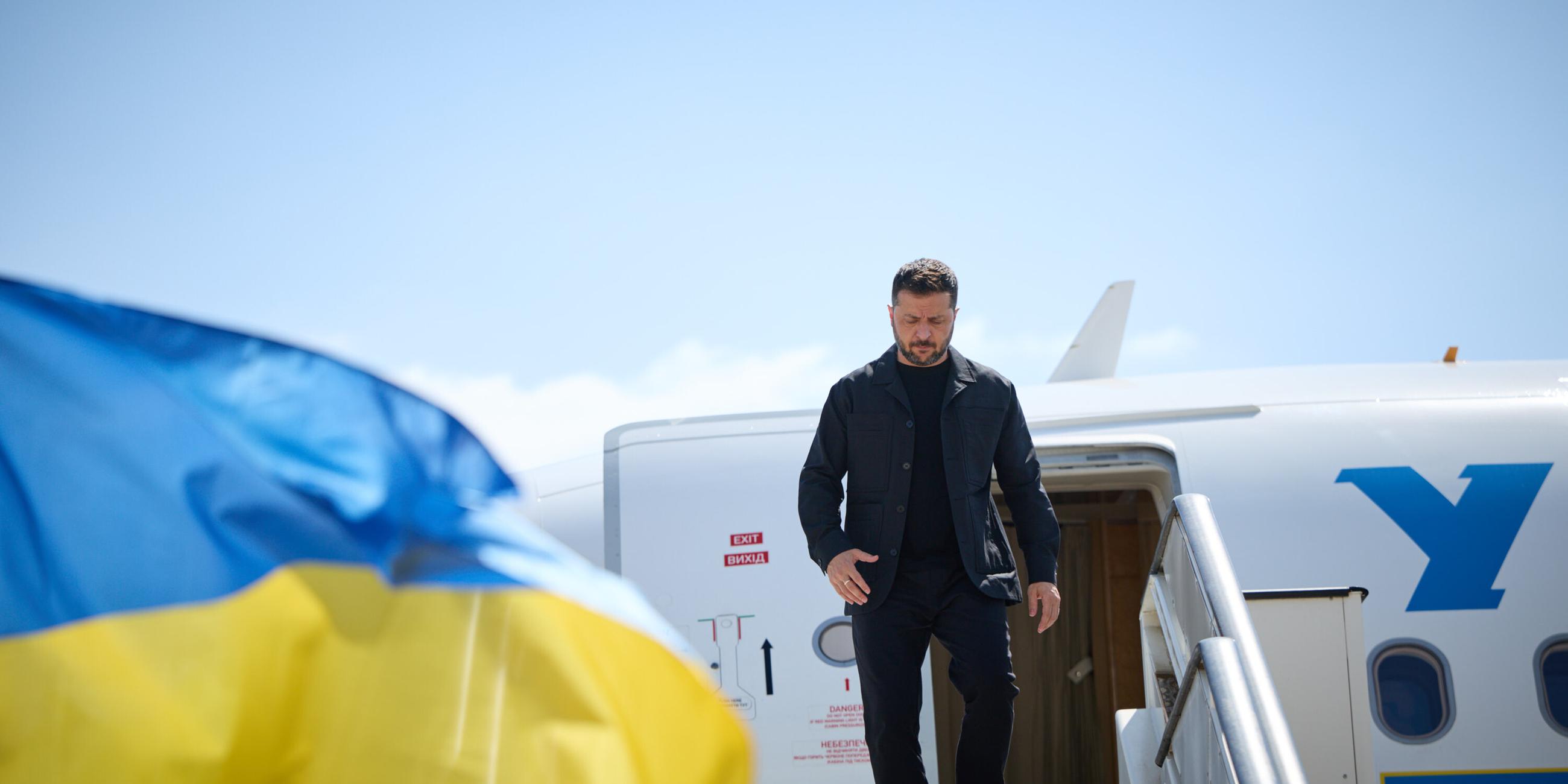 Selenskyj steigt aus einem Flugzeug aus. Links im Bild ist eine wehende ukrainische Flagge.
