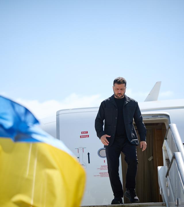 Selenskyj steigt aus einem Flugzeug aus. Links im Bild ist eine wehende ukrainische Flagge.