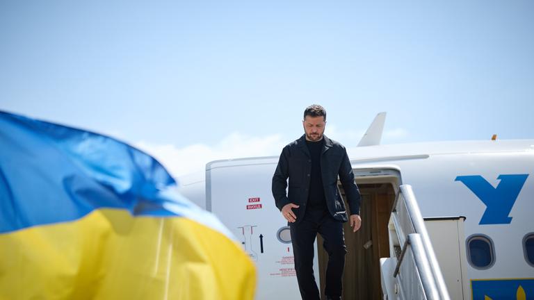 Selenskyj steigt aus einem Flugzeug aus. Links im Bild ist eine wehende ukrainische Flagge.