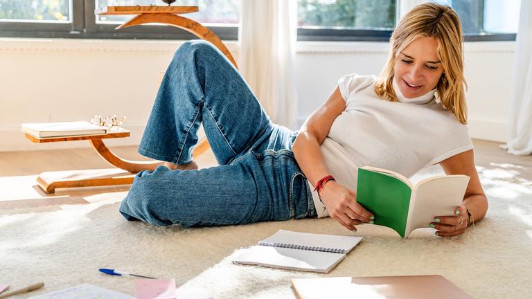 Frau liegt auf dem Boden und hält ein Buch in der Hand, um sie herum liegen Arbeitsmaterialen.