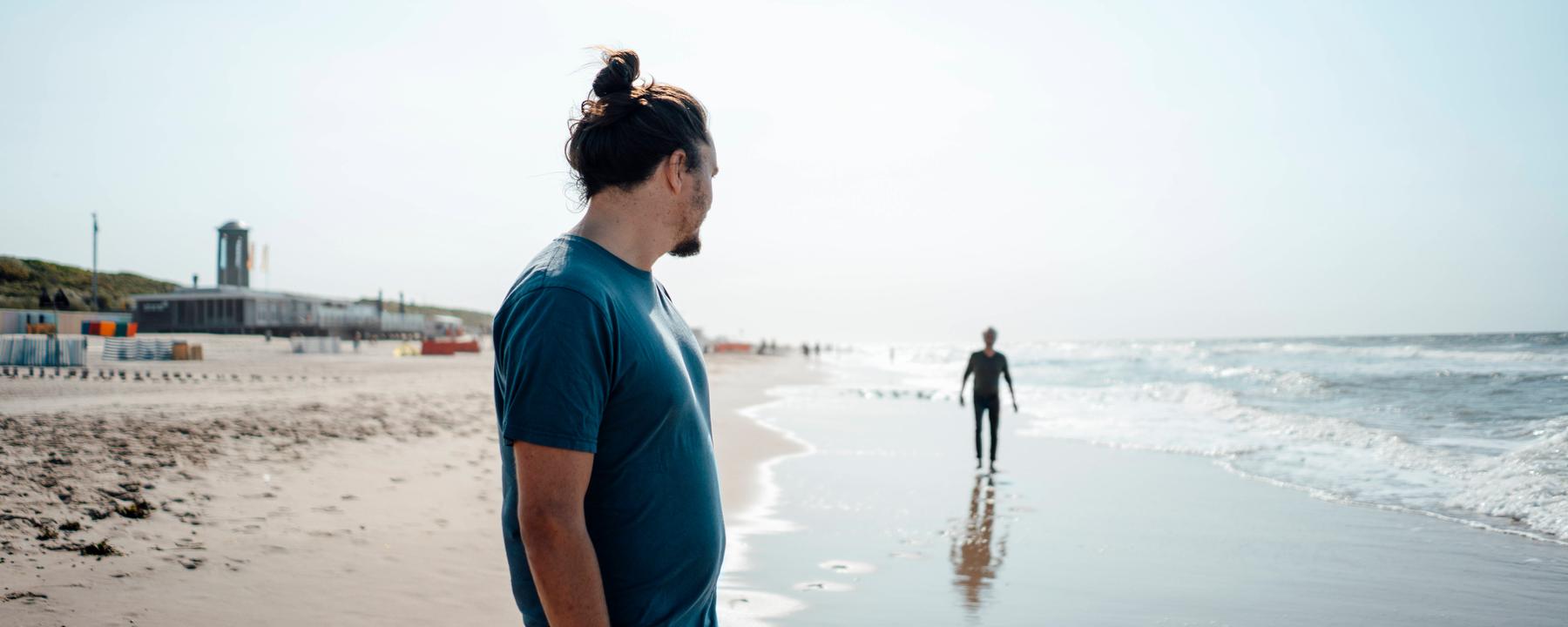Ein erwachsener Sohn steht an einem Strand und schaut zu seinem Vater in der Ferne.