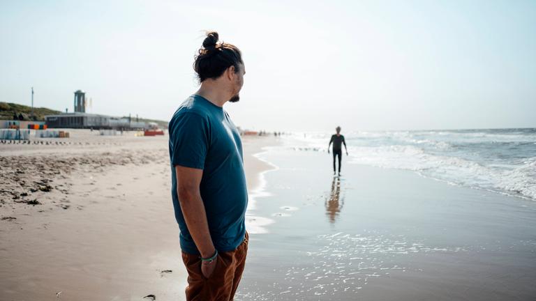 Ein erwachsener Sohn steht an einem Strand und schaut zu seinem Vater in der Ferne.