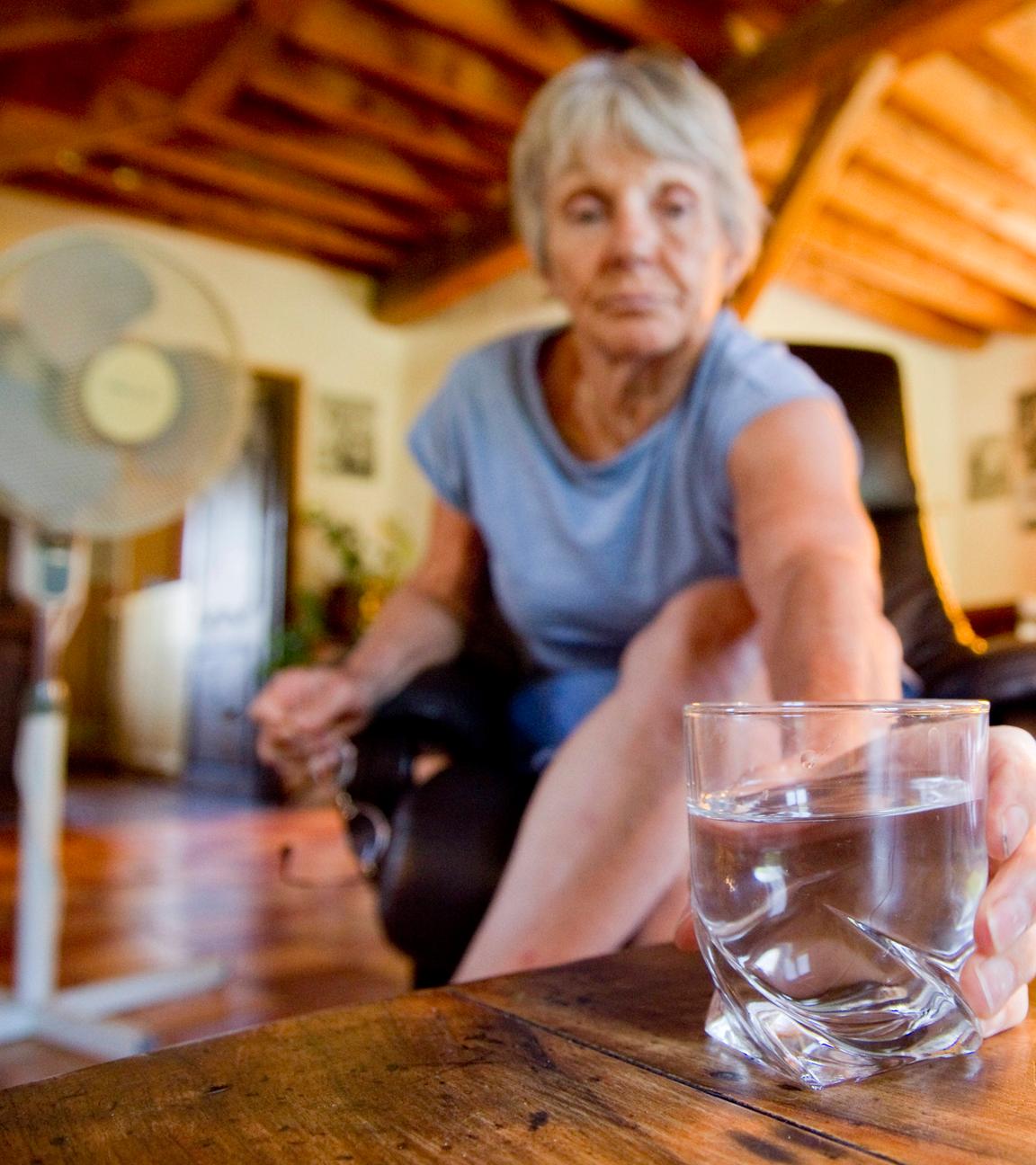 Ein Seniorin in ihrer Wohnung mit Ventilator im Hintergrund greift zu einem Wasserglas neben einer Wasserkaraffe.