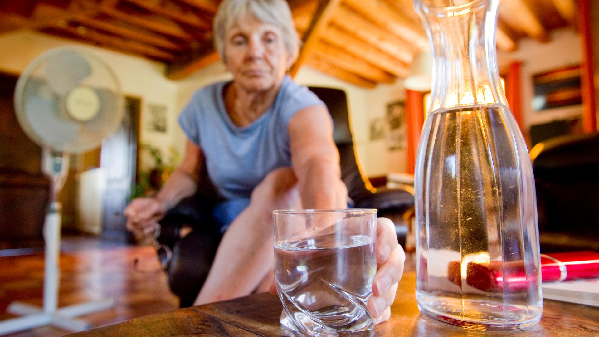 Ein Seniorin in ihrer Wohnung mit Ventilator im Hintergrund greift zu einem Wasserglas neben einer Wasserkaraffe.