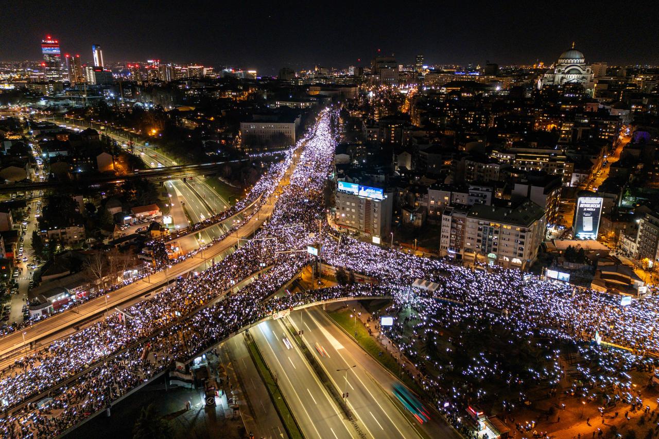 Eine große Menschenmasse blockiert abends eine Autobahnbrücke in Serbien. Ihre Handlyichter scheinen hell im Dunklen.