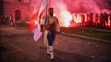 Ein Mann in traditioneller Kleidung geht, während regierungskritische Demonstranten Leuchtraketen werfen und Zäune umstoßen und die Bereitschaftspolizei verhöhnen.