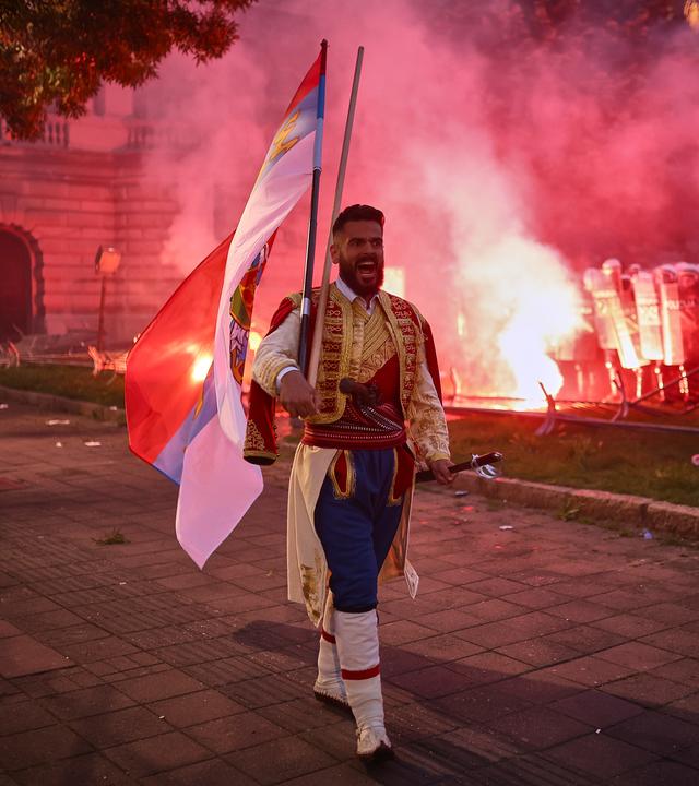 Ein Mann in traditioneller Kleidung geht, während regierungskritische Demonstranten Leuchtraketen werfen und Zäune umstoßen und die Bereitschaftspolizei verhöhnen.