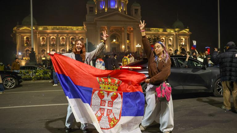 Zwei Frauen posieren mit der serbischen Flagge vor dem Parlament in Belgrad. Hinter ihnen feiern viele weitere Menschen den Wahlsieg der Studentenbewegung.