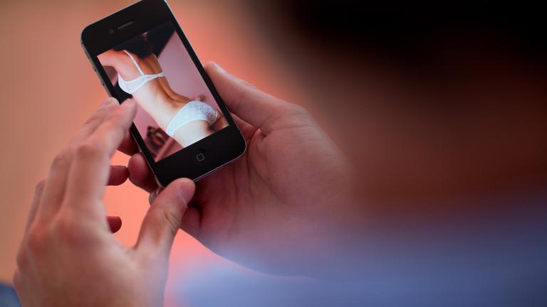 Ein junger Mann hält ein Smartphone, auf dem ein erotisches Foto einer jungen erwachsenen Frau zu sehen ist.