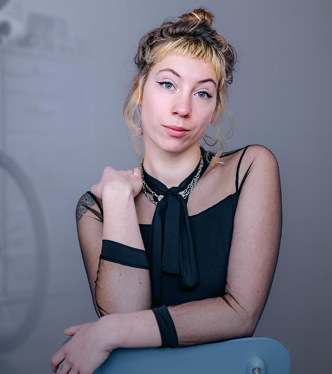 Pamina ist im Vordergrund zu sehen. Sie hat eine Hand auf einer Stuhllehne aufgelegt und die andere Hand hält sie auf ihre linke Schulter. Im Hintergrund ist ein Rollstuhl zu sehen. 