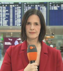 ZDF-Korrespondentin Brigitte Saar hält ein Mikrofon, im Hintergrund ist der Münchener Flughafen zu sehen.