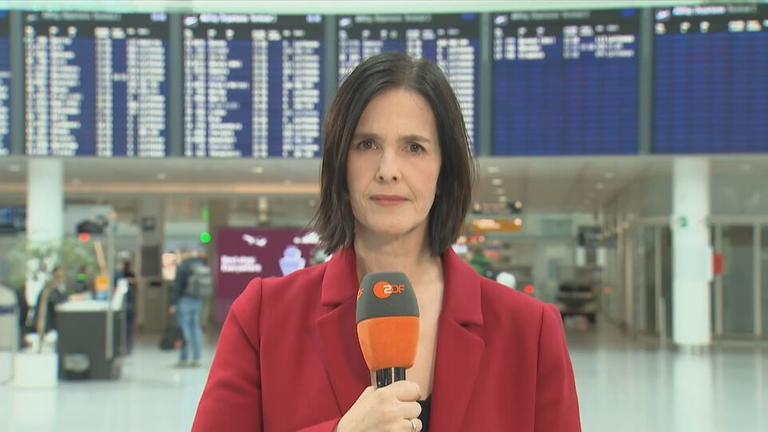 ZDF-Korrespondentin Brigitte Saar hält ein Mikrofon, im Hintergrund ist der Münchener Flughafen zu sehen.