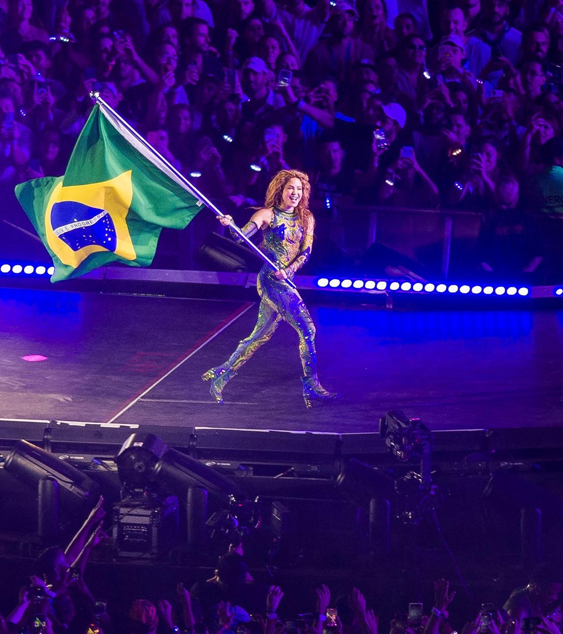 Sängerin Shakira läuft mit einer Brasilien-Flagge über eine Bühne, im Hintergrund Publikum.