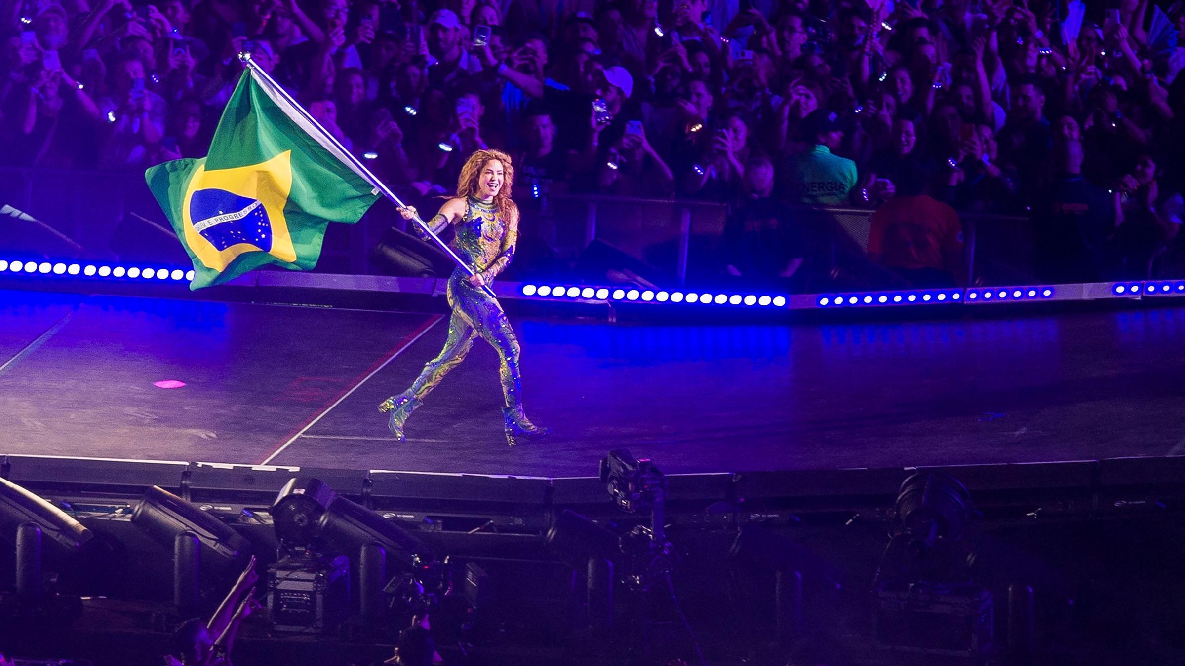 Sängerin Shakira läuft mit einer Brasilien-Flagge über eine Bühne, im Hintergrund Publikum.
