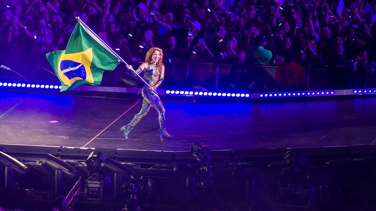 Sängerin Shakira läuft mit einer Brasilien-Flagge über eine Bühne, im Hintergrund Publikum.