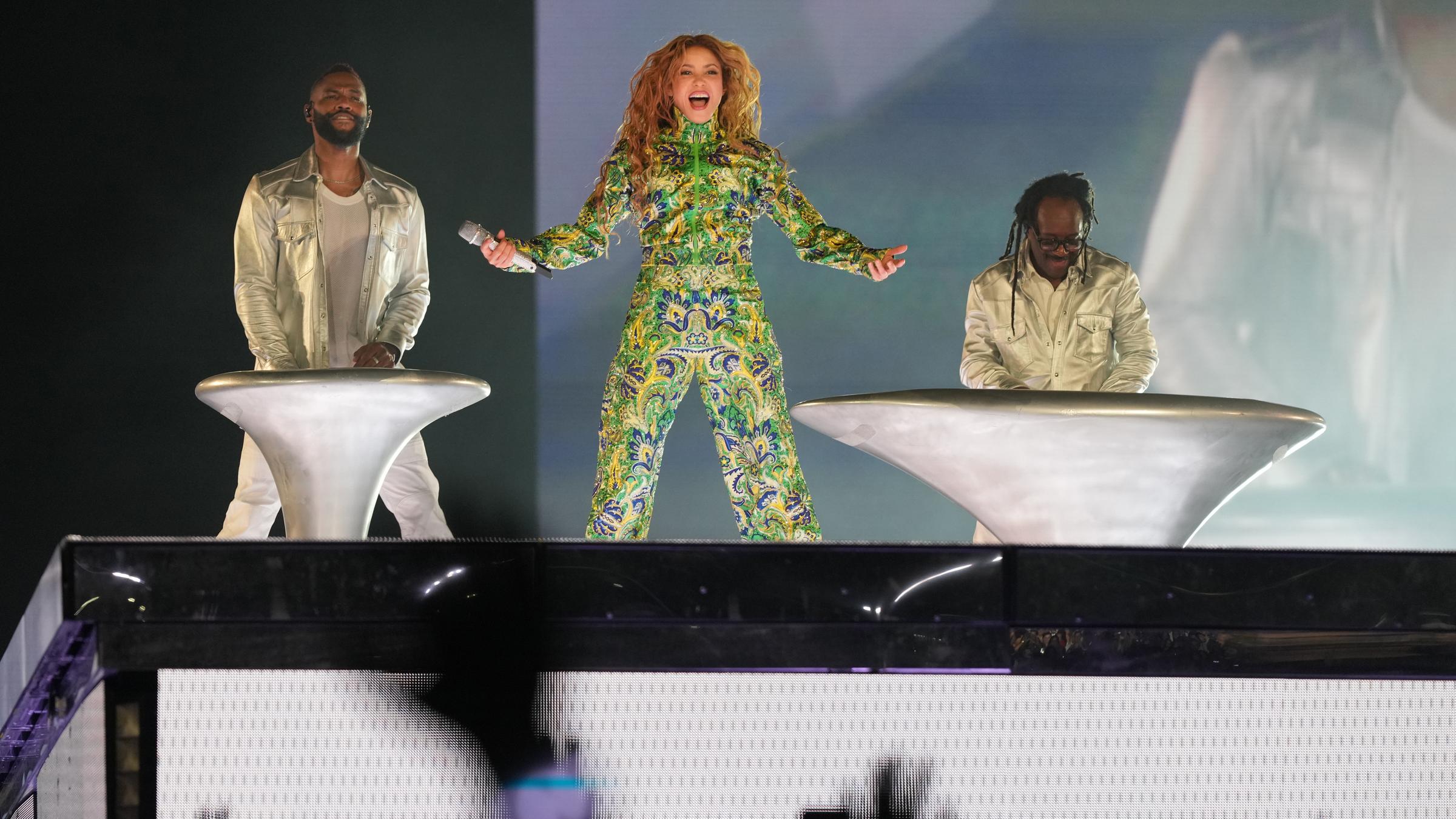 Sängerin Shakira performt in einer Jacke in den Nationalfarben Brasiliens - grün und gelb - auf der Bühne. Neben ihr stehen links und rechts zwei Männer.