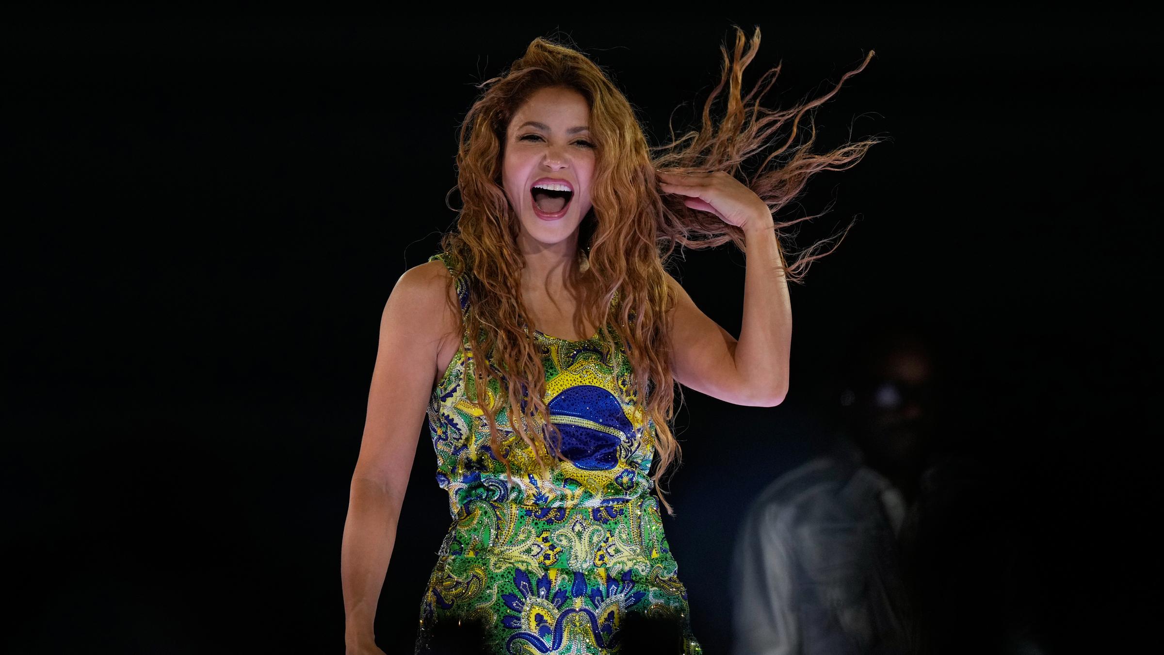 Sängerin Shakira steht lachend auf einer Bühne, sie trägt ein Kleid in grün, gelb und blau - den Nationalfarben Brasiliens.